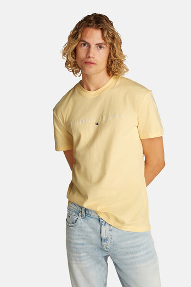 T-shirt jaune à manches courtes de TOMMY JEANS, avec logo sur le devant et porté avec des jeans bleus.