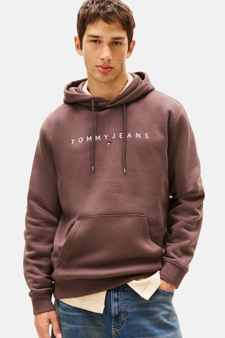 Bruine sweater met kap van TOMMY JEANS, met trekkoordjes en een buidelzak, gedragen over een lichtgekleurd overhemd.

