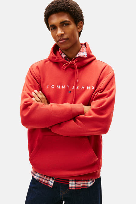 Rode sweater met kap van TOMMY JEANS, met gekruiste armen en een overhemd met ruitpatroon eronder.