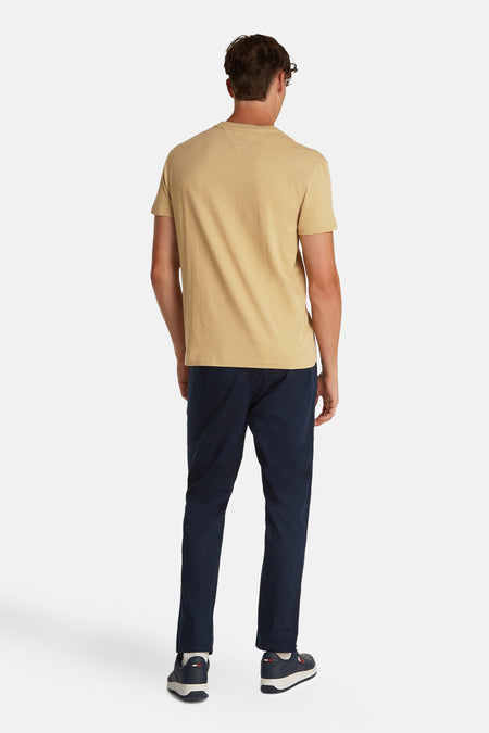 Beige T-shirt met korte mouwen van TOMMY JEANS, met effen kleur en rugaanzicht, gecombineerd met donkerblauwe broek en sneakers.