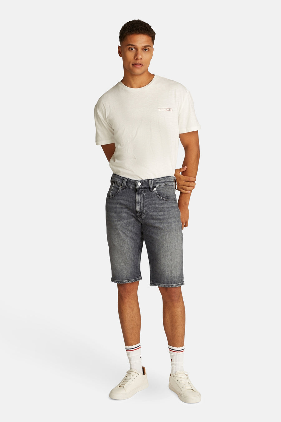Shorts en denim noir de TOMMY JEANS, portés avec un T-shirt blanc, des chaussettes blanches à rayures et des sneakers blanches.