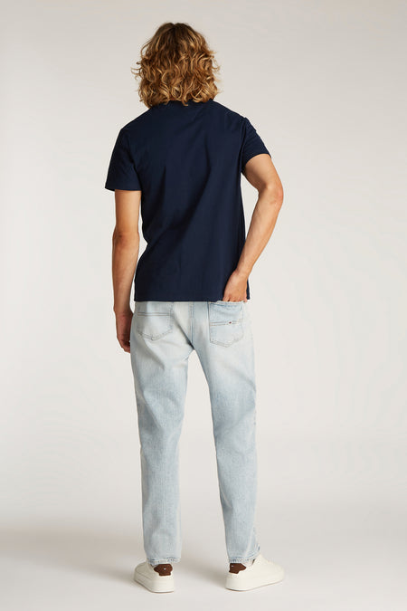 Blauw T-shirt met korte mouwen van TOMMY JEANS, met een man in lichte jeans en witte sneakers.
