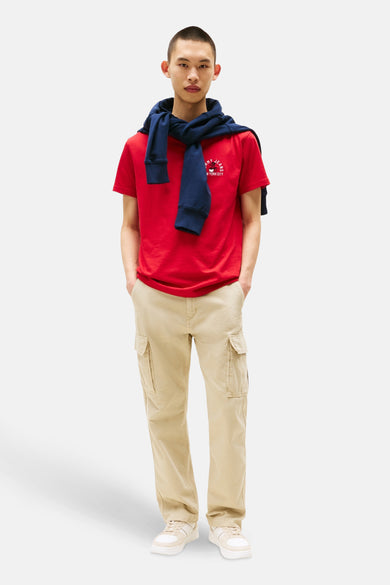 T-shirt rouge à manches courtes de TOMMY JEANS, associé nonchementlement un pull bleu marine sur les épaules, combiné avec un pantalon cargo beige et des baskets blanches.