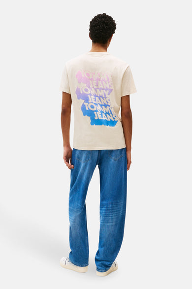 T-shirt beige à manches courtes de TOMMY JEANS, avec un dégradé de couleurs du logo 'Tommy Jeans' sur le dos, associé à des jeans décontractés et des baskets blanches.