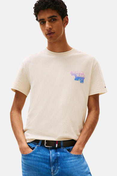 T-shirt beige à manches courtes de TOMMY JEANS, avec un imprimé sur la poitrine, porté avec un jean et une ceinture.