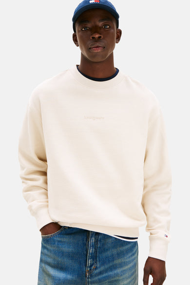Beige sweater van TOMMY JEANS, met wafeltextuur, ronde hals en lange mouwen, gedragen met blauwe jeans en een marineblauwe pet.