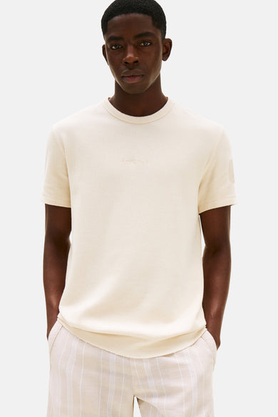 Beige T-shirt met korte mouwen van TOMMY JEANS, met een subtiele tekstprint op de borst en gedragen met gestreepte short.
