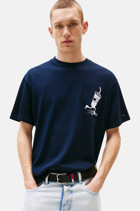 Blauw T-shirt met korte mouwen van TOMMY JEANS, met witte print van een beker op een skateboard, gedragen met een riem en lichte jeans.
