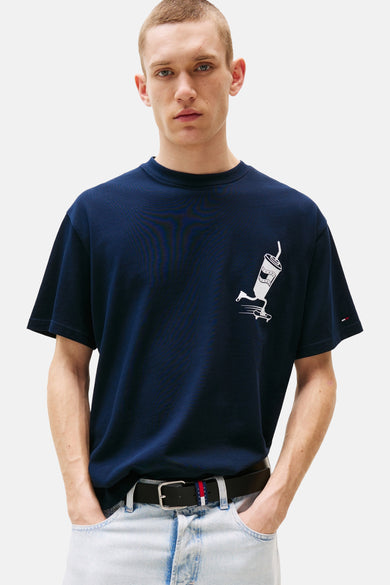 Blauw T-shirt met korte mouwen van TOMMY JEANS, met witte print van een beker op een skateboard, gedragen met een riem en lichte jeans.
