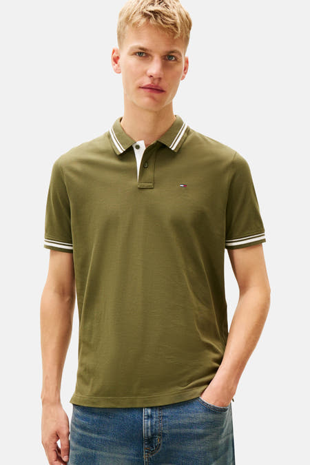 Kaki polo met korte mouwen van TOMMY JEANS, met witte strepen langs de kraag en mouwen, gedragen met jeans.
