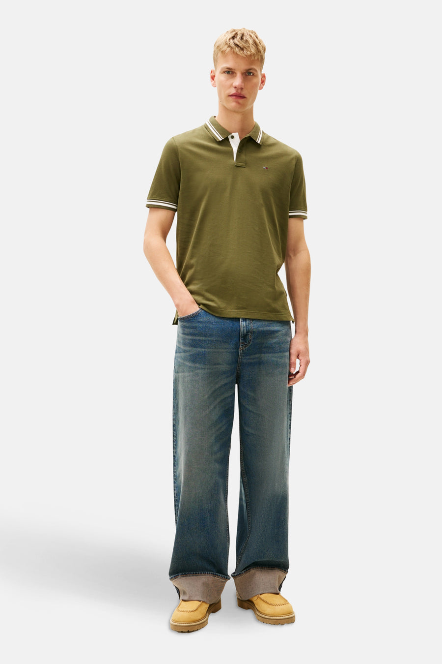 Man in kaki polo, wijde jeans en gele schoenen, staand.
