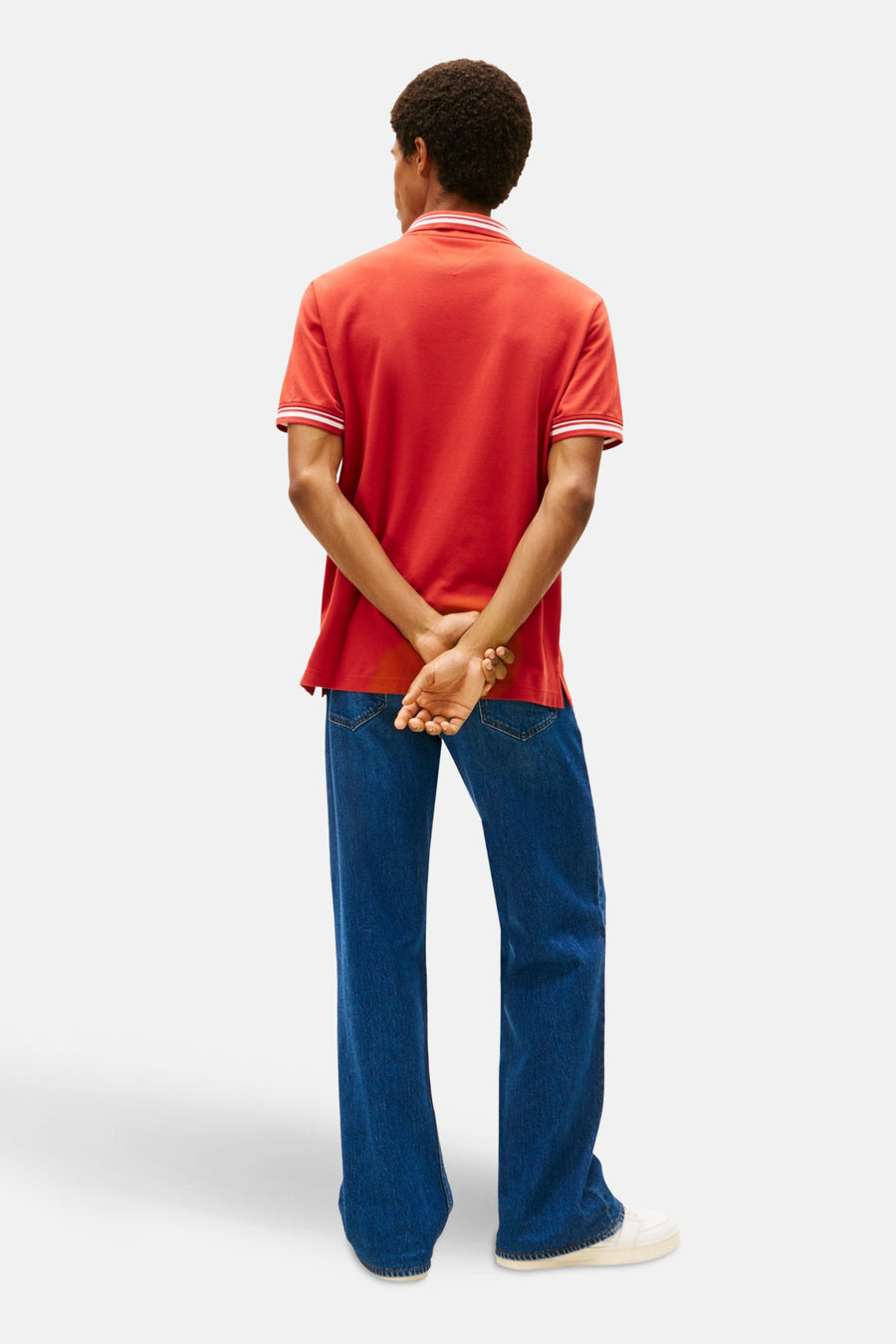 Man met gekruiste handen in een rode polo en blauwe spijkerbroek, van achteren gezien.