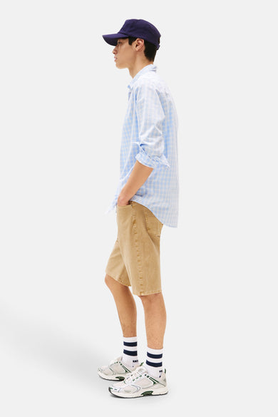 Beige jeansshort van TOMMY JEANS, met een opvallende textuur, gecombineerd met een blauw geruit overhemd en een donkerblauwe pet.
