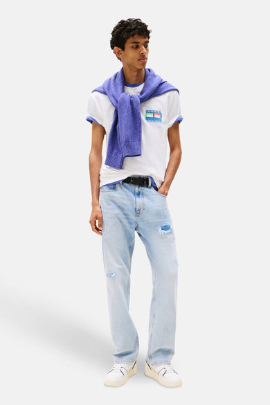 Wit T-shirt met korte mouwen van TOMMY JEANS, met logo op de borst, gecombineerd met een blauwe trui over de schouders en lichtblauwe jeans.
