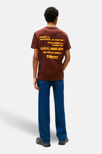 T-shirt brun à manches courtes de TOMMY JEANS, avec une impression textuelle jaune sur le dos, associé à un jean bleu.