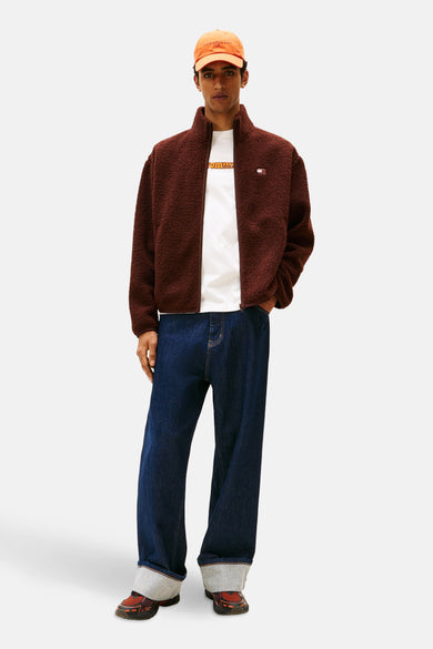 Bruine jas van TOMMY JEANS met rits, gedragen over een wit T-shirt en donkerblauwe jeans, met oranje pet.
