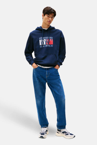 Blauwe sweater met kap van TOMMY JEANS, met een logo op de borst, gecombineerd met blauwe jeans en witte sneakers.
