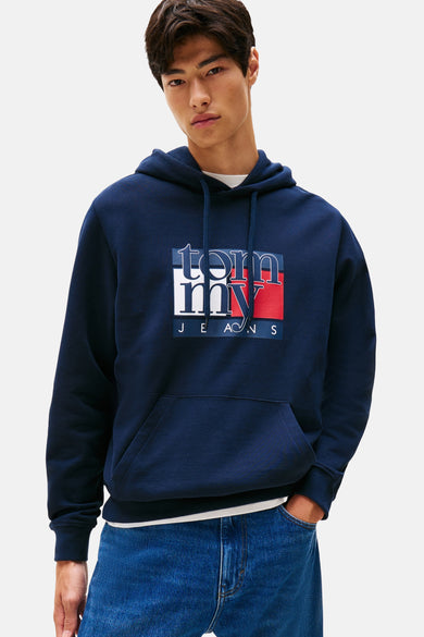 Blauwe sweater met kap van TOMMY JEANS, met logo op de voorkant, gedragen met een wit T-shirt en blauwe jeans.
