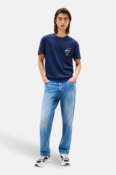 T-shirt bleu à manches courtes de Tommy Jeans, avec un logo de marque blanc imprimé sur la poitrine, porté avec des jeans bleus et des baskets blanches.