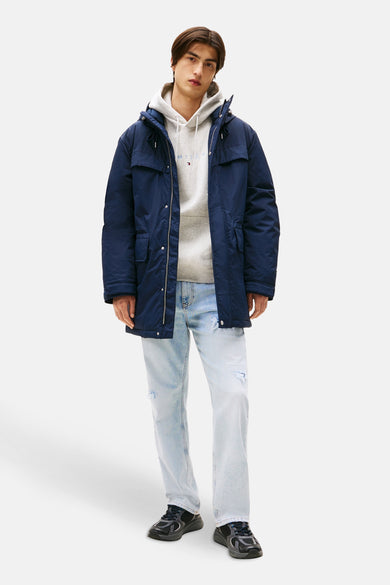 Man in blauwe jas, grijze hoodie en lichte jeans, staand tegen witte achtergrond.