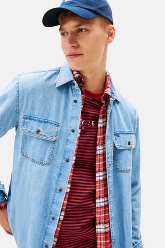 Jeanshemd in mid blue denim van TOMMY JEANS, gedragen over een geruit overhemd en een gestreept T-shirt.