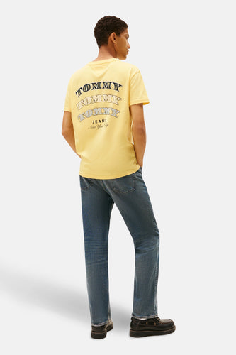 Man in gele T-shirt met Tommy Jeans-logo, blauwe jeans en zwarte schoenen.