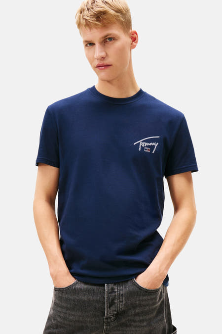 Blauw T-shirt met korte mouwen van TOMMY JEANS, met klein wit logo op de borst, gedragen met grijze jeans.
