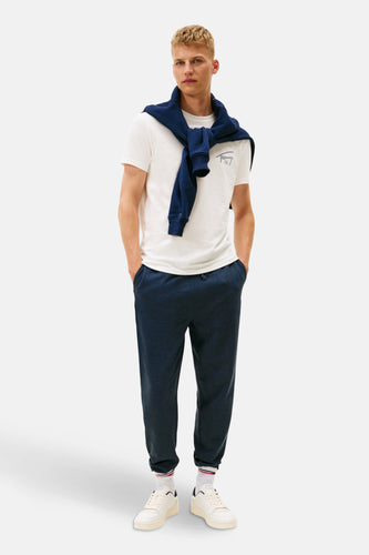 Man met witte T-shirt, blauwe trui, donkerblauwe joggingbroek en witte sneakers, staand.