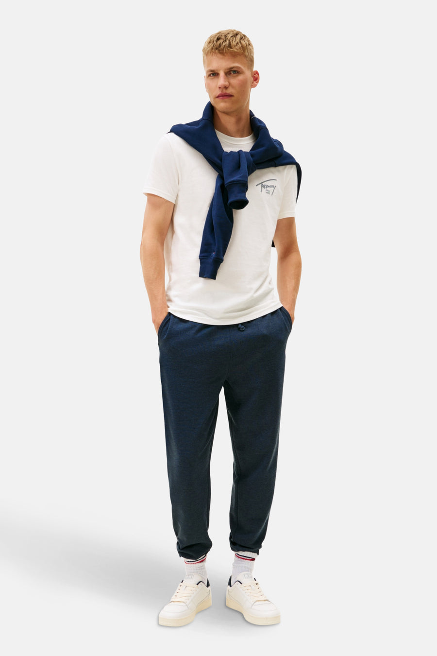 Man met witte T-shirt, blauwe trui, donkerblauwe joggingbroek en witte sneakers, staand.