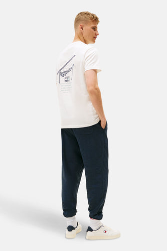 Een man staat met zijn handen in zijn zakken, gekleed in een wit T-shirt met een blauwe Tommy-print op de rug, een donkerblauwe joggingbroek en witte sneakers.