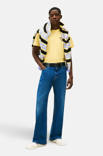 Geel T-shirt met korte mouwen van Tommy Jeans, gestyled met een gestreepte trui over de schouders en blauwe jeans.

