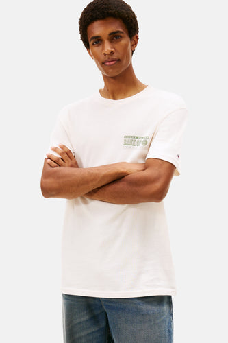 Man in een witte T-shirt met groene print en blauwe jeans, armen gekruist voor de borst, op een witte achtergrond.