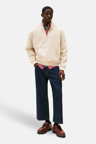 Man staand, beige trui, blauwe jeans, rode schoenen.