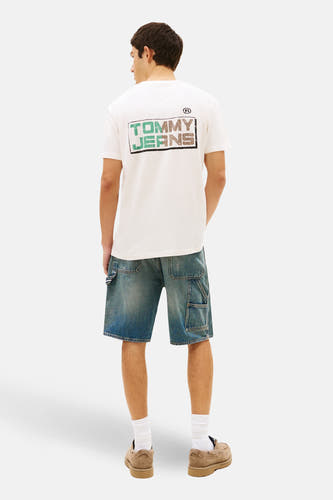 Ecru T-shirt met korte mouwen van TOMMY JEANS, met een grafische print op de rug, gecombineerd met blauwe denim shorts, witte sokken en suède schoenen.
