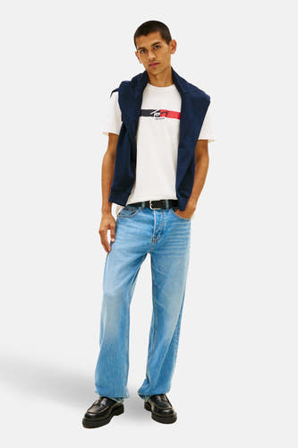 Wit T-shirt met korte mouwen van TOMMY JEANS, met marineblauwe sweater over de schouders en lichtblauwe jeans.