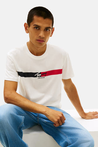 Wit T-shirt met korte mouwen van TOMMY JEANS, met rode en zwarte streep op de borst, gecombineerd met een blauwe jeansbroek.
