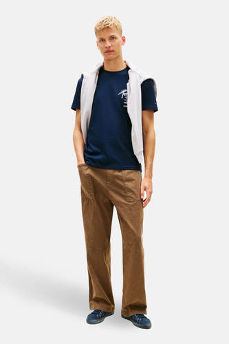 Blauw T-shirt met korte mouwen van TOMMY JEANS, met een sweater over de schouders en bruine broek.

