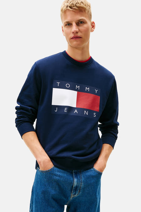 Blauwe sweater van TOMMY JEANS, met een groot logo op de borst, gecombineerd met een jeansbroek.
