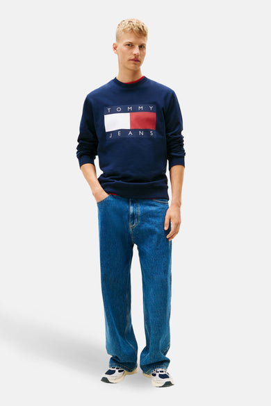 Blauwe sweater van TOMMY JEANS, met rechthoekig logo op de borst, gecombineerd met jeans en sneakers.
