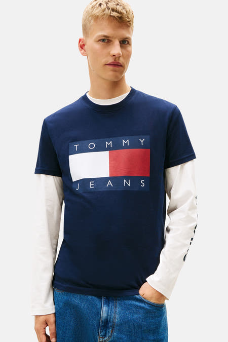 Blauw T-shirt met korte mouwen van TOMMY JEANS, gedragen onder een wit shirt met lange mouwen, met een grafisch logo op de voorkant.