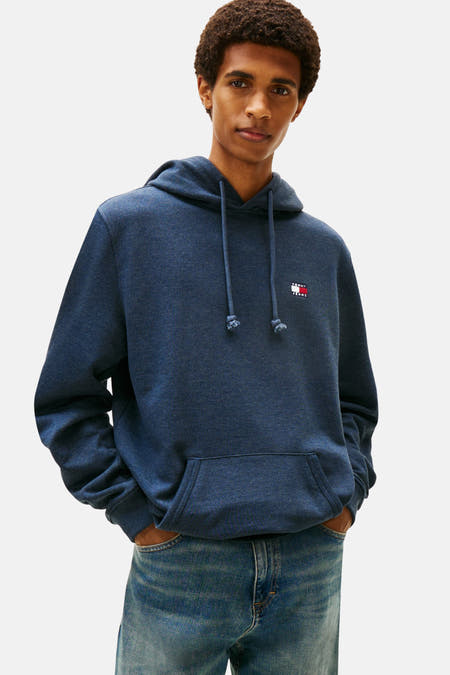 Blauwe sweater met kap van TOMMY JEANS, met trekkoorden en een buidelzak, gecombineerd met een jeansbroek.
