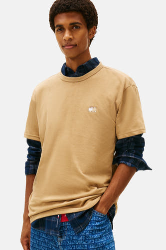Beige T-shirt met korte mouwen van TOMMY JEANS, gedragen over een blauw geruit overhemd en bedrukte jeans.
