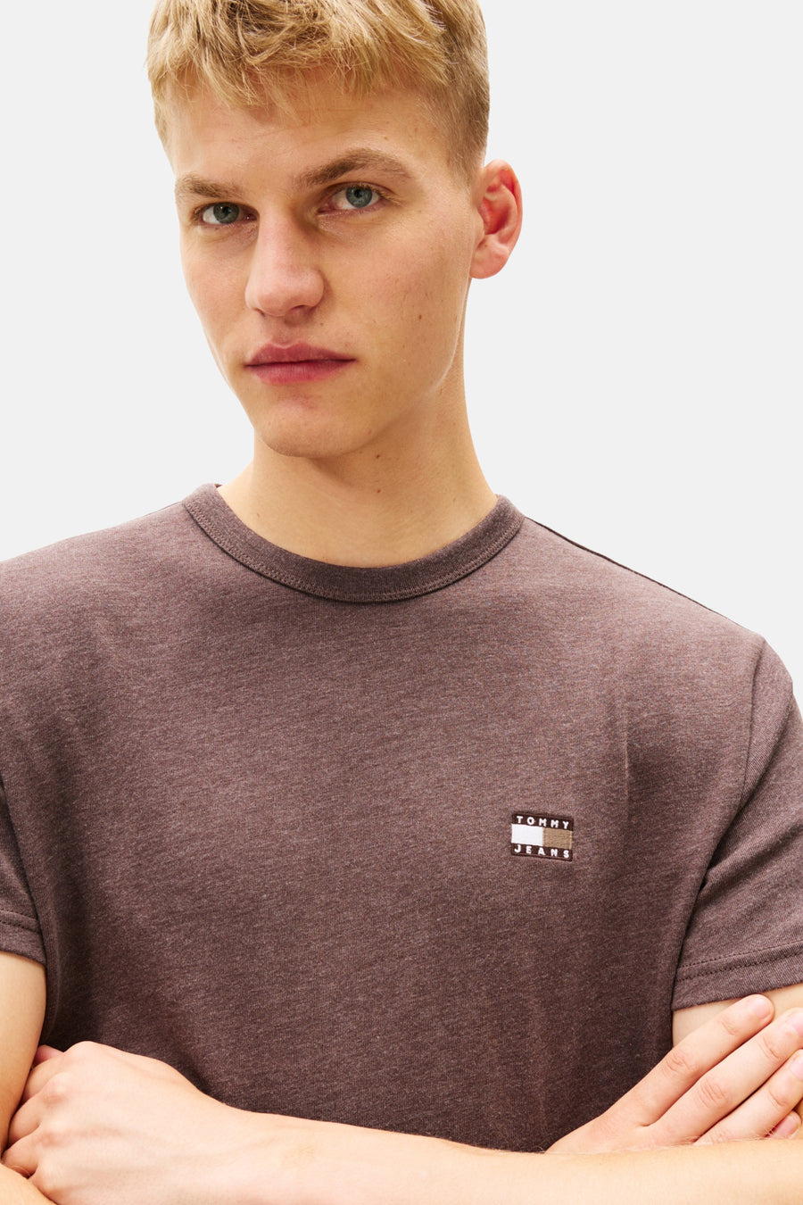 Man met blond haar draagt bruin Tommy Jeans T-shirt met gekruiste armen.