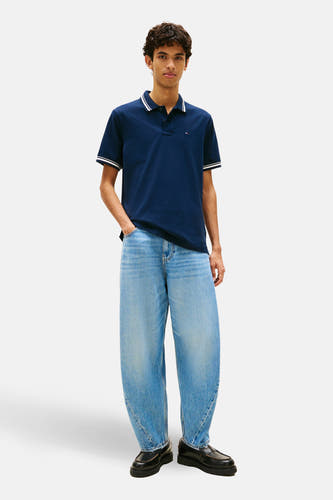 Blauwe polo met korte mouwen van TOMMY JEANS, met witte strepen op de kraag en mouwen, gecombineerd met wijde jeans en zwarte loafers.

