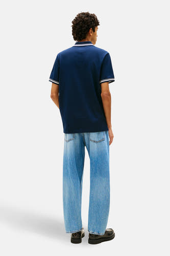 Blauwe polo met korte mouwen van TOMMY JEANS, met witte accenten op de kraag en mouwen. Gedragen met wijde jeans en zwarte schoenen.
