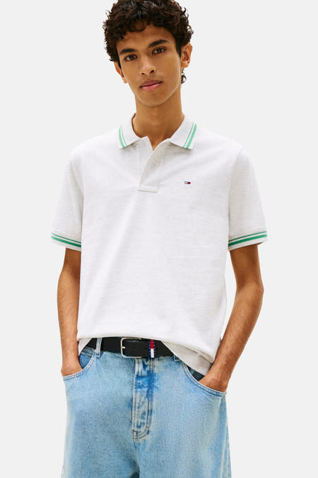 Grijs poloshirt met korte mouwen van TOMMY JEANS, afgewerkt met groene strepen aan de kraag en mouwen, gedragen met een zwarte riem en jeans.
