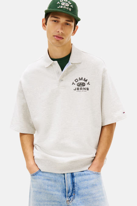 Grijze polo met korte mouwen van Tommy Jeans, met een logo op de borst, gedragen met een groene top en jeans.
