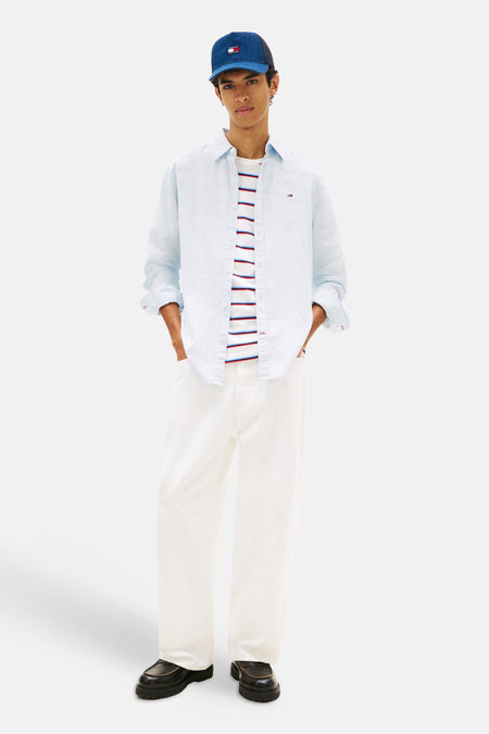 Blauw overhemd van TOMMY JEANS, gedragen over een gestreepte top, met witte broek en een blauwe pet.
