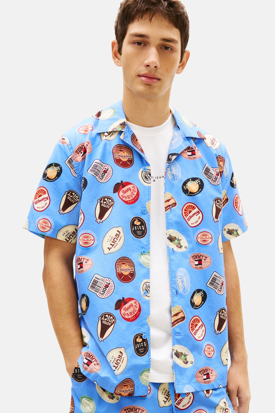 Blauw overhemd met korte mouwen van TOMMY JEANS, bedrukt met vintage fruitlogo's, gedragen over een wit T-shirt.
