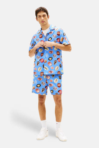 Blauw overhemd met korte mouwen van Tommy Jeans, voorzien van een herhaaldelijk etiketpatroon, gedragen met een wit T-shirt, shorts en sneakers.
